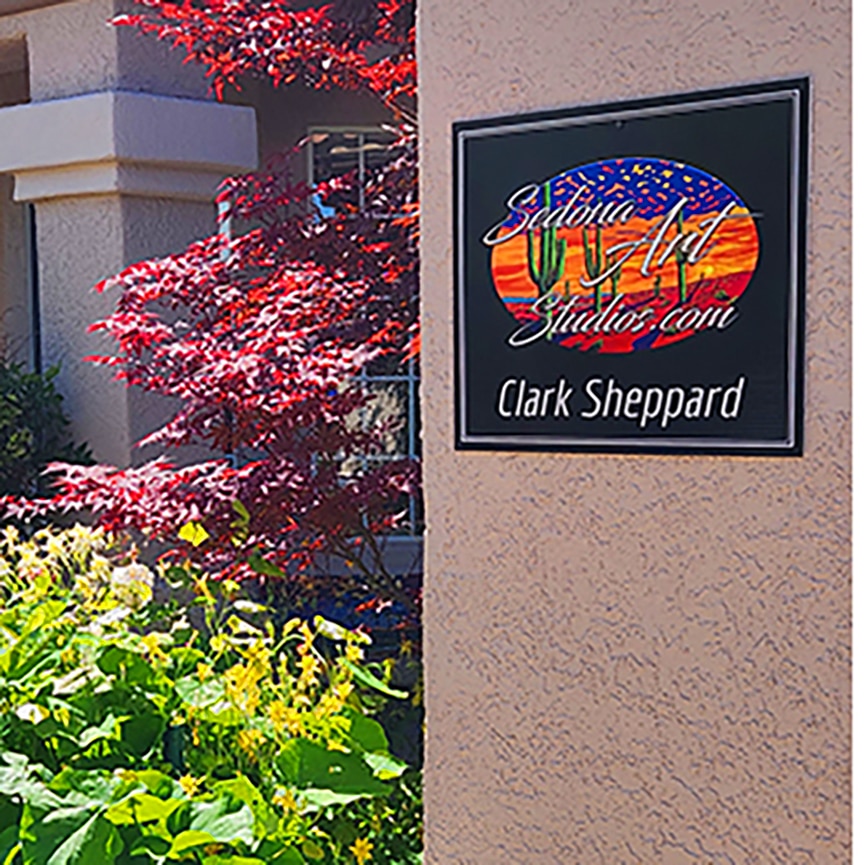 Sedona Art Studios Front Entrance Exterior view of a stucco wall with a colorful “SedonaArtStudios.com – Clark Sheppard” sign, framed by a red Japanese maple and yellow wildflowers in front of the studio.
