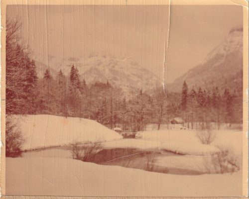 Vintage photograph of snowy valley with trees, small bridge, and mountains in the distance